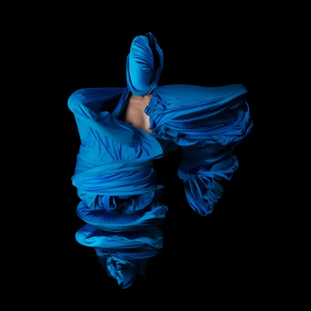 Performative photograph by Lizette Abraham: figure wrapped in fabrics on a black background, with blue fabric
