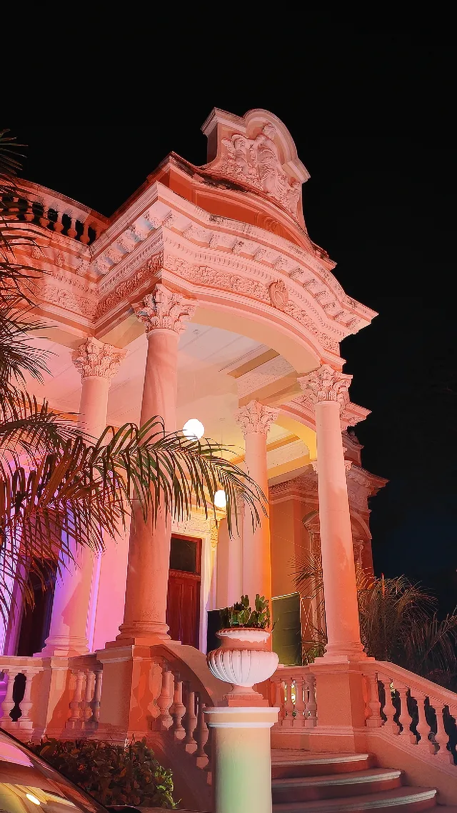 Pórtico de la fachada iluminado de noche durante un evento, con palmeras y balaustrada